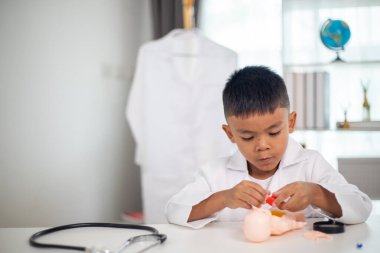 Genç bir çocuk doktorun ofisinde bir bebekle oynuyor. Oda steteskop ve oyuncak doktor seti de dahil olmak üzere tıbbi ekipmanlarla dolu. Çocuk beyaz bir laboratuvar önlüğü giyiyor.