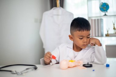 Genç bir çocuk doktorun ofisinde bir bebekle oynuyor. Oda steteskop ve oyuncak doktor seti de dahil olmak üzere tıbbi ekipmanlarla dolu. Çocuk beyaz bir laboratuvar önlüğü giyiyor.