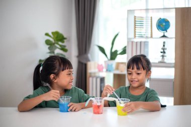 İki genç kız bir masada oturuyor, bir bilim deneyiyle oynuyorlar. Gülümsüyorlar ve eğleniyor gibiler.