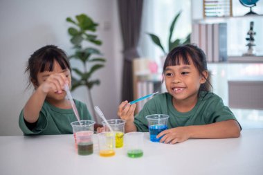 İki genç kız bir masada oturuyor, bir bilim deneyiyle oynuyorlar. Gülümsüyorlar ve eğleniyor gibiler.