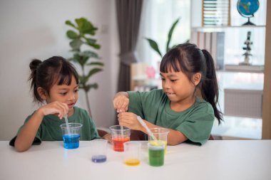 İki genç kız bir masada oturuyor, bir bilim deneyiyle oynuyorlar. Gülümsüyorlar ve eğleniyor gibiler.