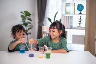 İki genç kız bir masada oturuyor, bir bilim deneyiyle oynuyorlar. Gülümsüyorlar ve eğleniyor gibiler.