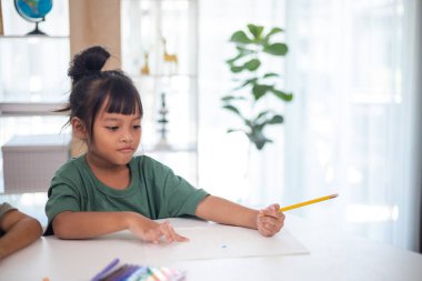 Genç bir kız elinde bir kalem ve bir kağıt parçasıyla bir masada oturuyor. Resim ve boyama pratiği yapıyor. Yeteneklerini evde geliştiriyor..