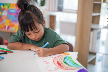 Genç bir kız elinde bir kalem ve bir kağıt parçasıyla bir masada oturuyor. Resim ve boyama pratiği yapıyor. Yeteneklerini evde geliştiriyor..