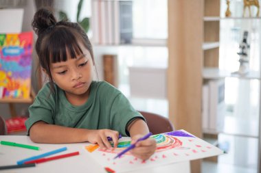 Genç bir kız elinde bir kalem ve bir kağıt parçasıyla bir masada oturuyor. Resim ve boyama pratiği yapıyor. Yeteneklerini evde geliştiriyor..