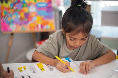 Genç bir kız elinde bir kalem ve bir kağıt parçasıyla bir masada oturuyor. Resim ve boyama pratiği yapıyor. Yeteneklerini evde geliştiriyor..