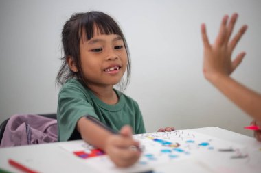 Genç bir kız elinde bir kalem ve bir kağıt parçasıyla bir masada oturuyor. Resim ve boyama pratiği yapıyor. Yeteneklerini evde geliştiriyor..