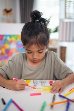 Genç bir kız elinde bir kalem ve bir kağıt parçasıyla bir masada oturuyor. Resim ve boyama pratiği yapıyor. Yeteneklerini evde geliştiriyor..