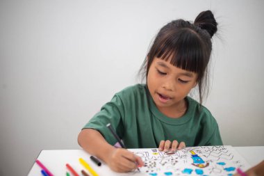 Genç bir kız elinde bir kalem ve bir kağıt parçasıyla bir masada oturuyor. Resim ve boyama pratiği yapıyor. Yeteneklerini evde geliştiriyor..