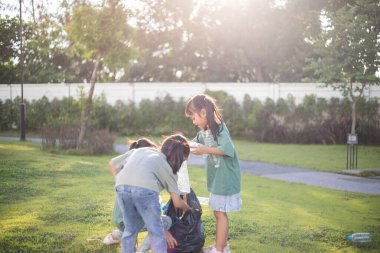 Üç genç kız bir parkta çöp topluyor. Ellerinde plastik şişeler ve bir çöp torbası var. Kızlar parkı temiz tutmak için harekete geçerken sahne olumlu ve umut verici.