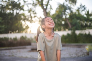 Genç bir kız parkta duruyor, gülümsüyor ve gülüyor. O yeşil bir gömlek giyiyor ve örgülü saçı var. Park yeşil çimen ve ağaçlarla dolu.
