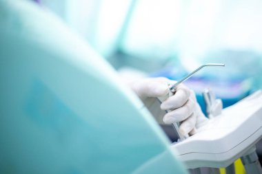 Bir dişçi beyaz ve yeşil bir odada diş aletini tutuyor. Oda temiz ve iyi aydınlatılmış, bir sandalye ve lavabo ile. Dişçi eldiven takıyor ve elindeki işe odaklanmış durumda.