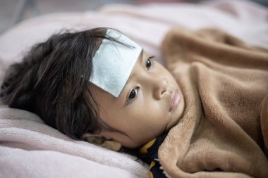 Alnı bandajlı bir çocuk yatağa uzanıyor. Çocuğun alnında bandaj var ve ateşi var.
