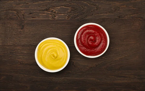 将两个红色番茄酱和黄色芥末的白色陶瓷碗放在褐色桌子的背景上 在正上方有复制空间 — 图库照片