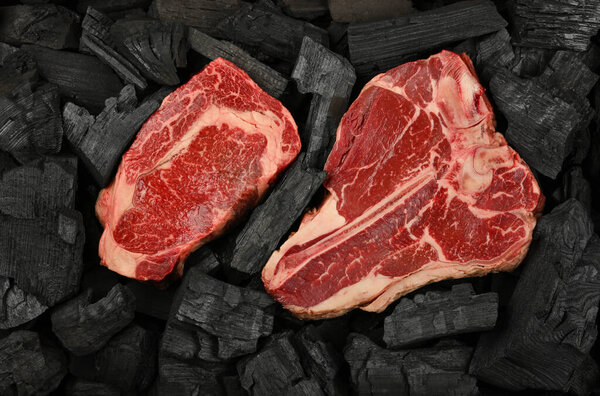 Close up one aged prime marbled raw ribeye and porterhouse T bone beef steaks on black lump charcoal pieces ready for barbecue grill cooking, elevated top view, directly above