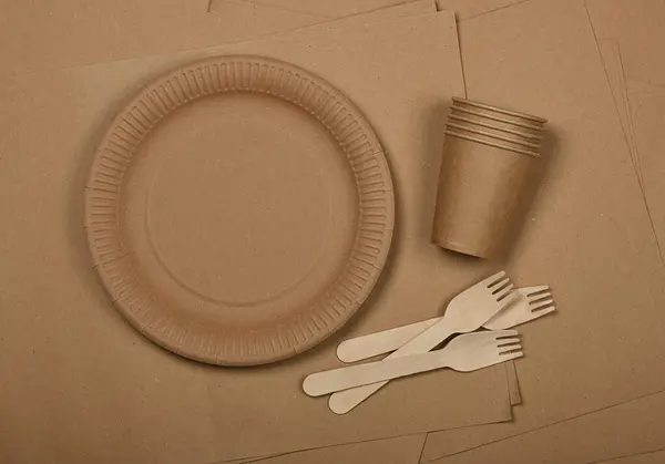 Close up disposable paper plates, takeaway coffee cups and wooden forks on brown paper background, natural eating and drinking utensils, close up, elevated table top view, directly above