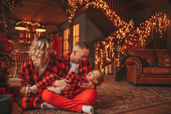 微笑积极的父母 带着小儿子 穿着红色格子睡衣等着圣诞老人回家 时尚的家庭喜欢在家里笑着拥抱亲吻 12月25日晚上庆祝新年花环和彩灯 — 图库照片