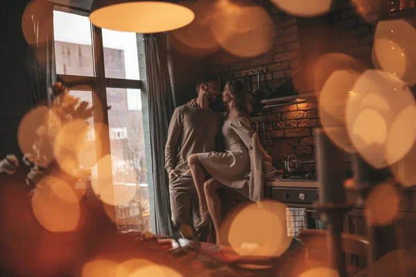 Portrait of young lovely couple hugging indoor eve 25 December. Lovers laughing hugs kisses waiting xmas at home. Celebrating new year garlands lights noel in elegant knitwear outfit tenderness