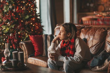 Young teen boy with long hair thoughtful look sad eyes negative mood angry and crying at home. Stylish zoomer gen Z pensive on new year holidays with xmas tree bokeh lights garlands eve 25 december clipart