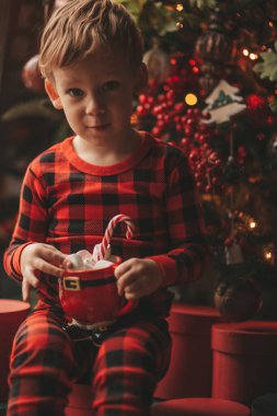 Mucizevi Noel Baba 'yı bekleyen mutlu küçük hayalperest çocuk renkli lolipopun tadını çıkarıyor. Kırmızı kareli uykucu neşeli çocuk yeni yılı kutluyor Noel ağacının yanında marshmallowlu Xmas bardağı tutuyor 25 Aralık arifesi