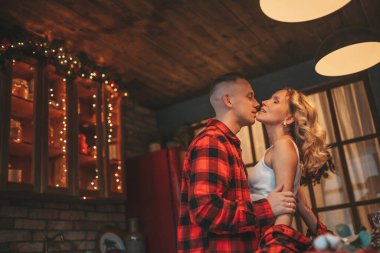 25 Aralık arifesinde evde kucaklaşan güzel bir çiftin portresi. Aşıklar gülüyor, öpücükler evde mucize bekliyor. Yeni yılı kutlamak için çelenk ışıkları kırmızı pijamalar içinde Noel Baba 'nın ihtirası.