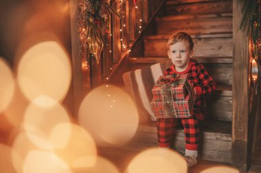 Gülümse küçük çocuk Noel Baba 'nın hediyelerini açmasını beklerken eğleniyor. Kırmızı kareli uykucu neşeli çocuk yeni yılı hediye kutularıyla kutluyor çelenk yakıyor Noel arifesinde 25 Aralık 'ta