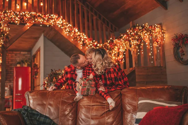 微笑积极的父母 带着小儿子 穿着红色格子睡衣等着圣诞老人回家 时尚的家庭喜欢在家里笑着拥抱亲吻 12月25日晚上庆祝新年花环和彩灯 — 图库照片
