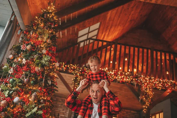 笑顔の小さな男の子がお互いの屋内待っている奇跡と楽しんで父親の首に座っています ノエルの木の冬の季節のガーランドライトで新年を祝うクリスマスの精神前夜25 12月父方の愛 ロイヤリティフリーのストック画像