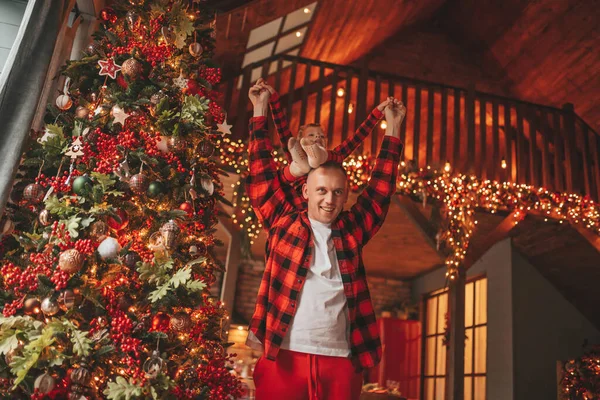 笑顔の小さな男の子がお互いの屋内待っている奇跡と楽しんで父親の首に座っています ノエルの木の冬の季節のガーランドライトで新年を祝うクリスマスの精神前夜25 12月父方の愛 ストック写真