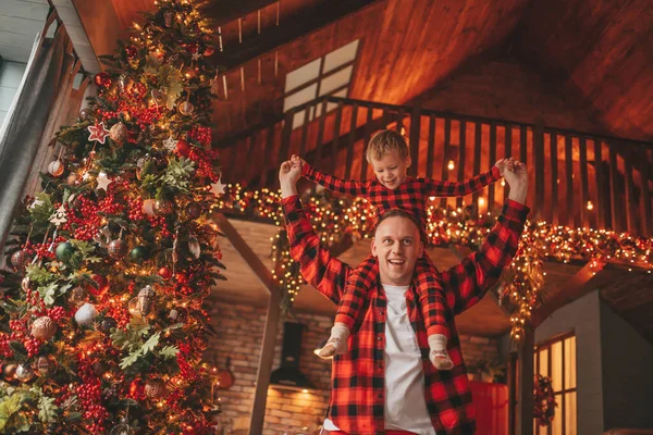 笑顔の小さな男の子がお互いの屋内待っている奇跡と楽しんで父親の首に座っています ノエルの木の冬の季節のガーランドライトで新年を祝うクリスマスの精神前夜25 12月父方の愛 ロイヤリティフリーのストック写真