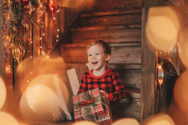 Gülümse küçük çocuk Noel Baba 'nın hediyelerini açmasını beklerken eğleniyor. Kırmızı kareli uykucu neşeli çocuk yeni yılı hediye kutularıyla kutluyor çelenk yakıyor Noel arifesinde 25 Aralık 'ta