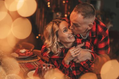 25 Aralık arifesinde evde kucaklaşan güzel bir çiftin portresi. Aşıklar gülüyor, öpücükler evde mucize bekliyor. Yeni yılı kutlamak için çelenk ışıkları kırmızı pijamalar içinde Noel Baba 'nın ihtirası.