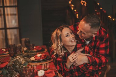 25 Aralık arifesinde evde kucaklaşan güzel bir çiftin portresi. Aşıklar gülüyor, öpücükler evde mucize bekliyor. Yeni yılı kutlamak için çelenk ışıkları kırmızı pijamalar içinde Noel Baba 'nın ihtirası.