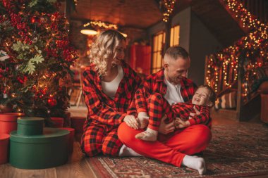 Smile active parents with small son in red checkered sleepwears waiting Santa indoor. Stylish family love laughing hugs kisses at home. Celebrate New year garlands and lights eve 25 December Noel tree clipart