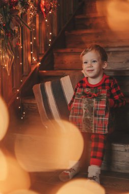 Gülümse küçük çocuk Noel Baba 'nın hediyelerini açmasını beklerken eğleniyor. Kırmızı kareli uykucu neşeli çocuk yeni yılı hediye kutularıyla kutluyor çelenk yakıyor Noel arifesinde 25 Aralık 'ta