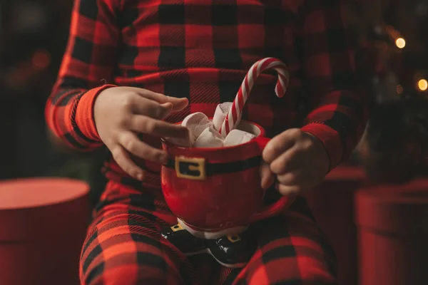 stock image Happy little boy dreamer waiting at miracle Santa enjoy sweets color lollipop. Red checkered sleepwear cheerful kid celebrating new year hold Xmas cup with marshmallows near noel tree eve 25 december