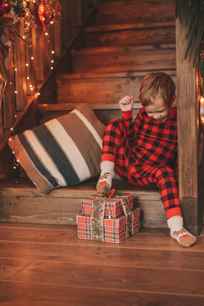Smile small child being active having fun waiting for miracle Santa open presents. Red checkered sleepwear cheerful kid celebrating new year with gifts boxes garlands lights noel at eve 25 december