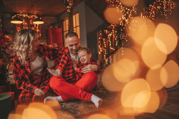 赤い小さな息子を持つ笑顔活動的な両親は サンタの屋内を待っているチェッカー付きの眠りにつく スタイリッシュな家族の愛笑い抱擁自宅でキス 新年のガーランドとライトを祝う12月25日ノエルの木 — ストック写真