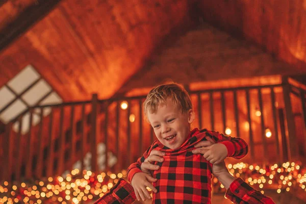 笑顔の小さな男の子がお互いの屋内待っている奇跡と楽しんで父親の首に座っています ノエルの木の冬の季節のガーランドライトで新年を祝うクリスマスの精神前夜25 12月父方の愛 ストックフォト