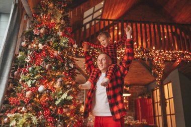 Gülümse küçük çocuk, babanın boynuna oturup içeride eğleniyor ve mucize bekliyor. Noel ağacı kış sezonunun çelenk ışıklarıyla yeni yılı kutluyoruz Noel ruhu 25 Aralık baba sevgisi arifesi