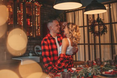 25 Aralık arifesinde evde kucaklaşan güzel bir çiftin portresi. Aşıklar gülüyor, öpücükler evde mucize bekliyor. Yeni yılı kutlamak için çelenk ışıkları kırmızı pijamalar içinde Noel Baba 'nın ihtirası.