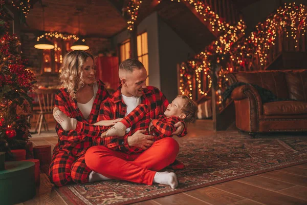 赤い小さな息子を持つ笑顔活動的な両親は サンタの屋内を待っているチェッカー付きの眠りにつく スタイリッシュな家族の愛笑い抱擁自宅でキス 新年のガーランドとライトを祝う12月25日ノエルの木 — ストック写真