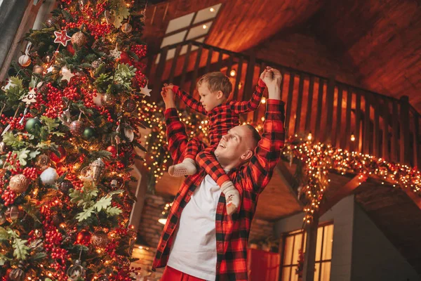 笑顔の小さな男の子がお互いの屋内待っている奇跡と楽しんで父親の首に座っています ノエルの木の冬の季節のガーランドライトで新年を祝うクリスマスの精神前夜25 12月父方の愛 ストック画像