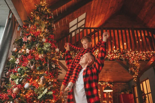 笑顔の小さな男の子がお互いの屋内待っている奇跡と楽しんで父親の首に座っています ノエルの木の冬の季節のガーランドライトで新年を祝うクリスマスの精神前夜25 12月父方の愛 ストック写真