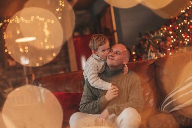 Gülümse küçük çocuk ve baba içeride Noel Baba 'yı beklerken kucaklaşıp öpüşüyorlar. Noel ağacı kış sezonunun çelenk ışıklarıyla yeni yılı kutluyoruz Noel ruhu 25 Aralık baba sevgisi arifesi