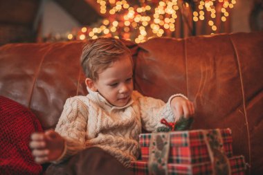 Gülümse küçük çocuk Noel Baba 'nın hediyelerini açmasını beklerken eğleniyor. Yeni yılı hediye kutularıyla, çelenklerle kutlayan sıradan örgü kıyafetli neşeli çocuk Noel arifesi 25 Aralık.