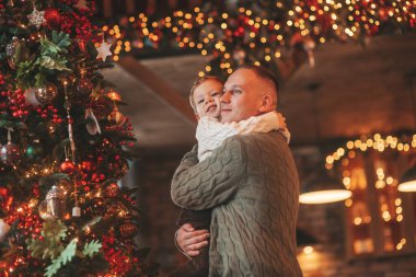 Gülümse küçük çocuk ve baba içeride Noel Baba 'yı beklerken kucaklaşıp öpüşüyorlar. Noel ağacı kış sezonunun çelenk ışıklarıyla yeni yılı kutluyoruz Noel ruhu 25 Aralık baba sevgisi arifesi