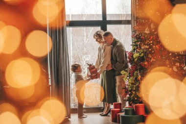 Evde Noel Baba 'yı bekleyen örgü bej kazaklı mutlu aile portresi. Gülümseyen ebeveynler küçük oğlunu kucaklıyor ve öpüyor. 25 Aralık, Noel ağacı hediyeleriyle Noel 'i kutluyor.