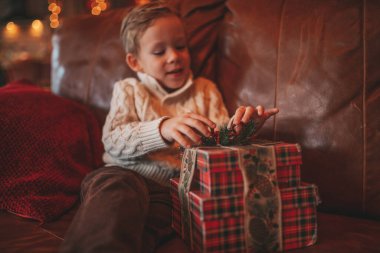 Gülümse küçük çocuk Noel Baba 'nın hediyelerini açmasını beklerken eğleniyor. Yeni yılı hediye kutularıyla, çelenklerle kutlayan sıradan örgü kıyafetli neşeli çocuk Noel arifesi 25 Aralık.