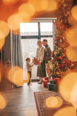 Evde Noel Baba 'yı bekleyen örgü bej kazaklı mutlu aile portresi. Gülümseyen ebeveynler küçük oğlunu kucaklıyor ve öpüyor. 25 Aralık, Noel ağacı hediyeleriyle Noel 'i kutluyor.